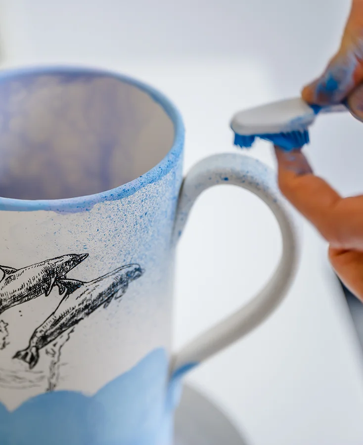Eine Hand benutzt eine Zahnbürste, um blaue Farbe auf eine weiße Keramiktasse zu spritzen, die mit einer Zeichnung von Delphinen verziert ist.
