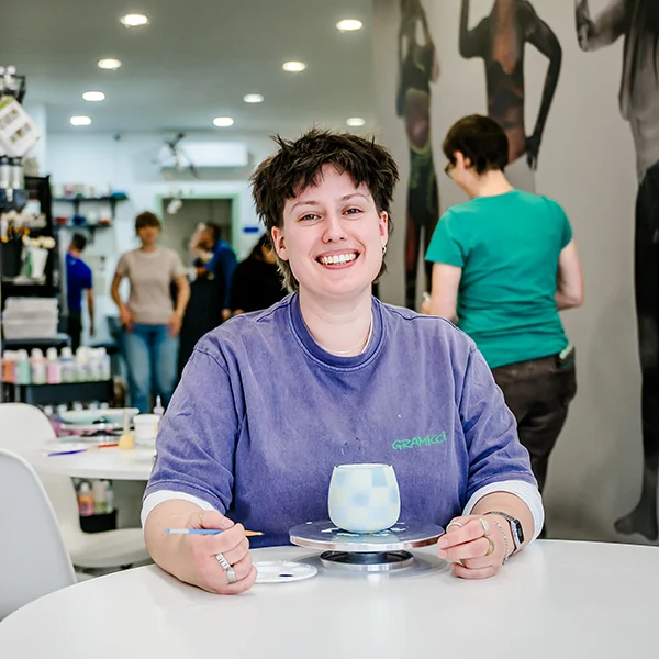Eine Person mit kurzen dunklen Haaren, die ein lila Hemd trägt, sitzt an einem Tisch in einem Kunstatelier und lächelt in die Kamera. Sie bemalt gerade einen Keramikgegenstand, während im Hintergrund Kunstzubehör und andere Personen zu sehen sind.